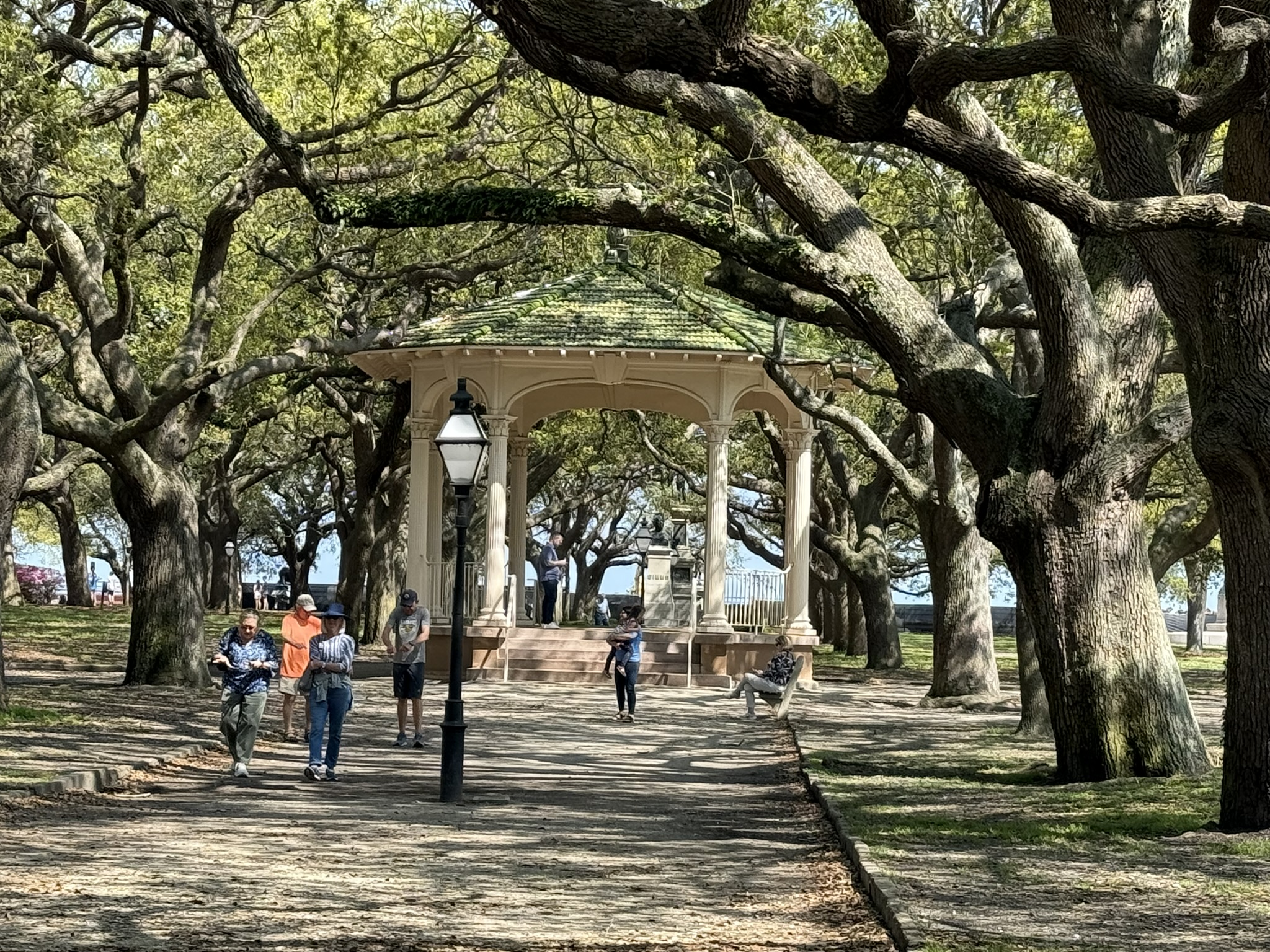 Gazebo in White Point Park with large oak trees surrounding it. You can see a light post and people in the picture and Charleston's bay in the background of one of the best things to do in Charleston