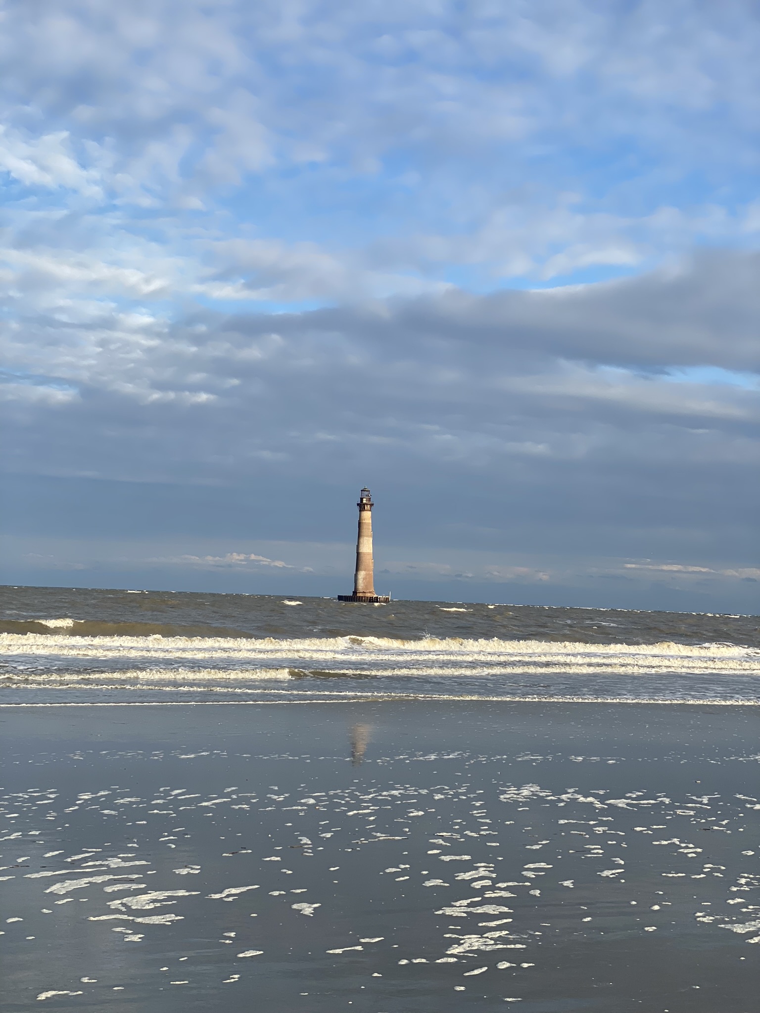 Ocean waves at the end of Folly Beach with a white and red lighthouse in the background. Blue sky with streaked white and grey clouds.
