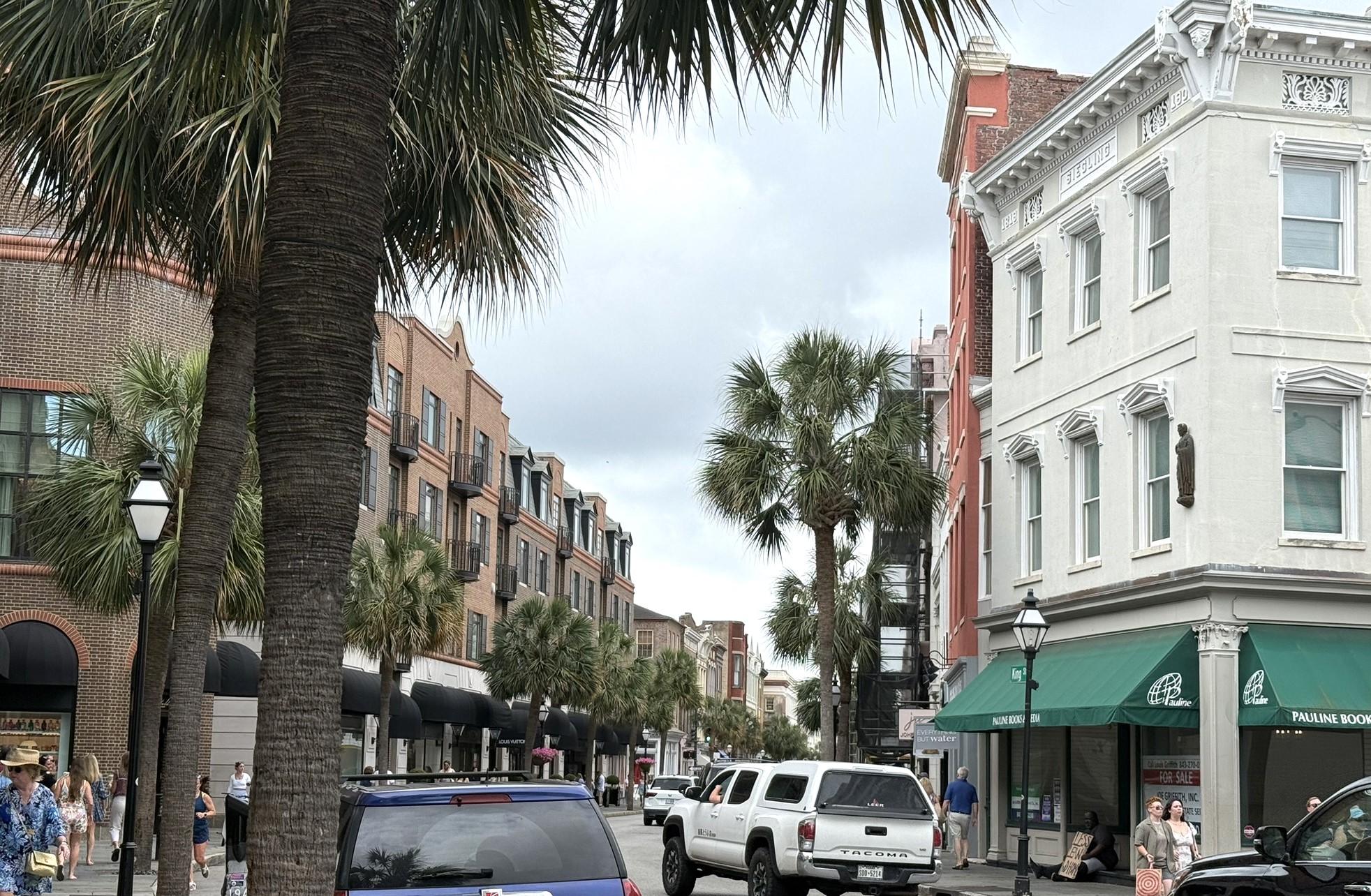 A view down King St in Charleston, SC. The street is lined with palmetto trees and shops on either side with people walking on the sidewalks and cars driving down the street