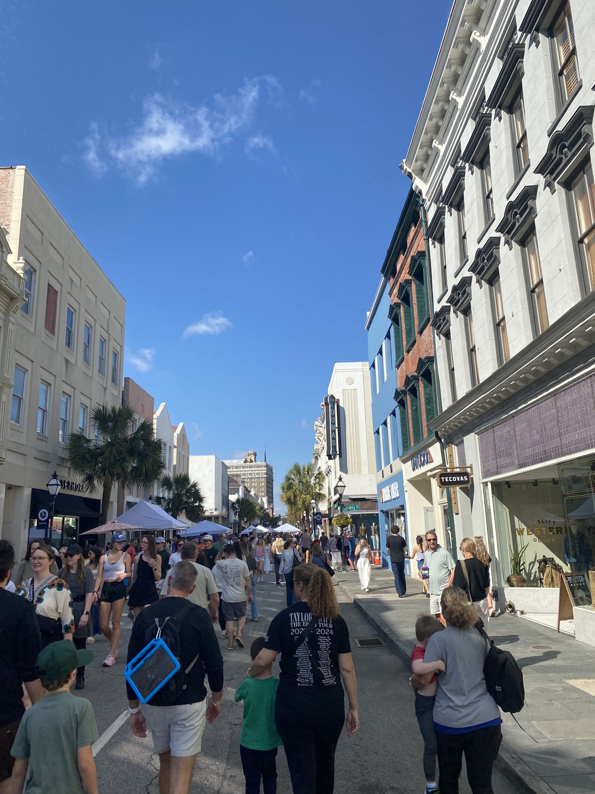Pedestrians walking on a crowded street with classic Charlestonian buildings on either side. Blue sky with several small fluffy white clouds
