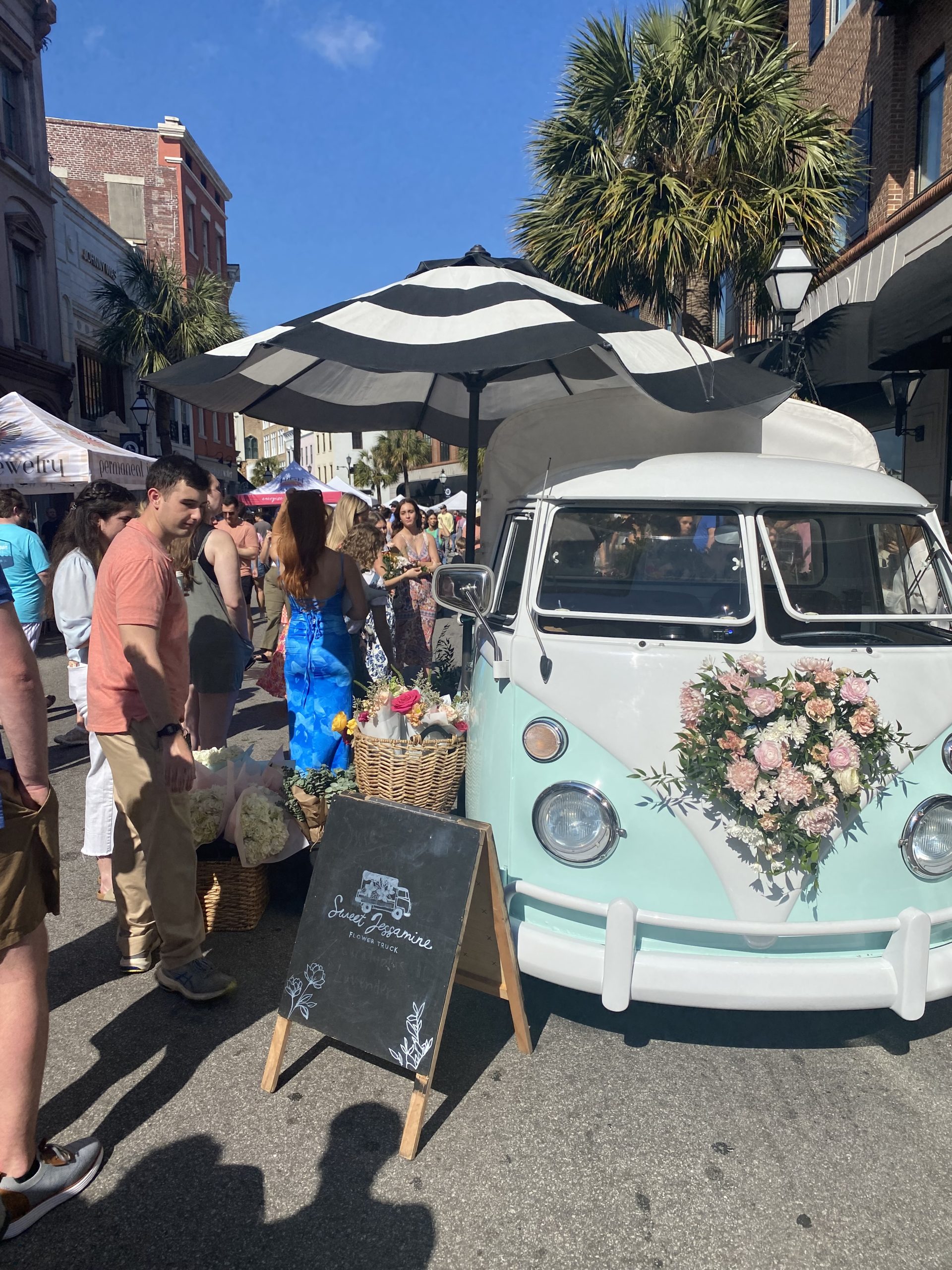 Light teal and white classic VW bus with black and white umbrella shading fresh cut flowers for sale on side of bus. Line of people waiting to purchase flowers