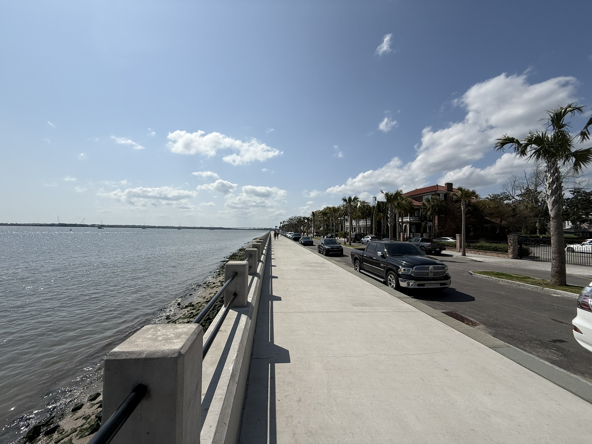 Sunny day with several white clouds in the blue sky. There is a walkway with the bay to the left and the street, palm trees and houses to the right. This is Charleston's Battery on Charleston's Itinerary for Couples
