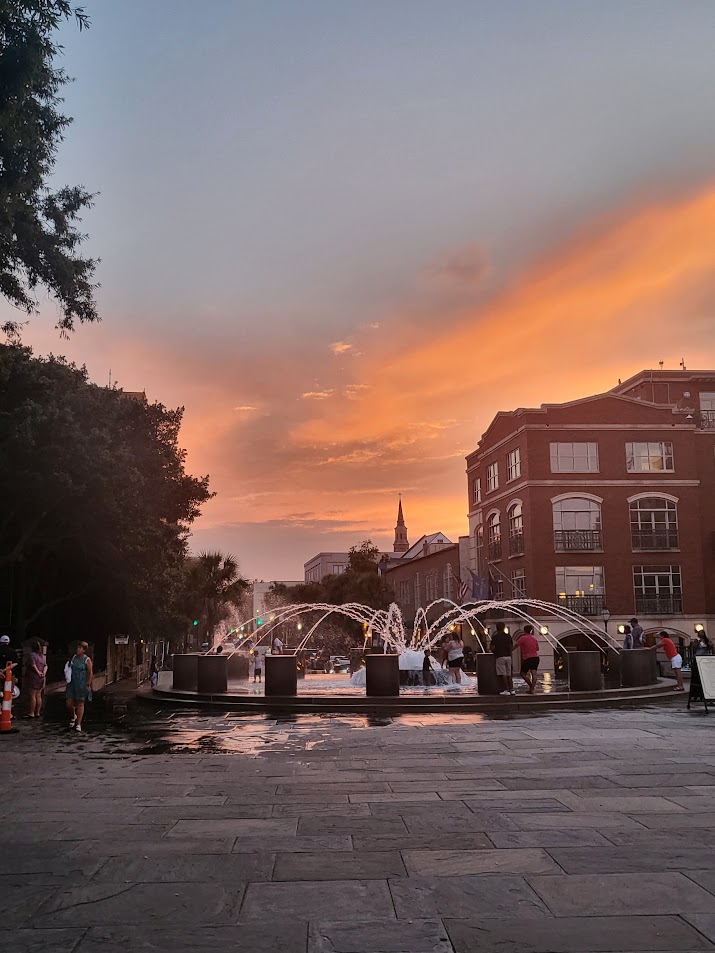 Fountain at the east end of Charleston's Waterfront park where you can see some trees and Charleston buildings in the background. The sun is setting in the sky