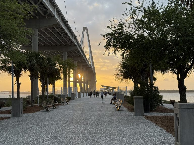 Large walkway lined with palm and cypress trees on either side. A large bridge is in front of you as well as water as the sun sets