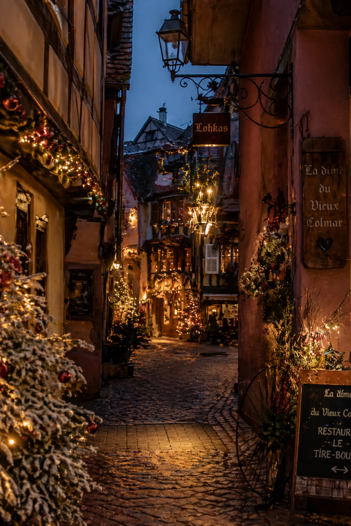 A fairy tale alleyway with snow covered Christmas trees and lights lite on both sides as it becomes dark in Colmar, France.
