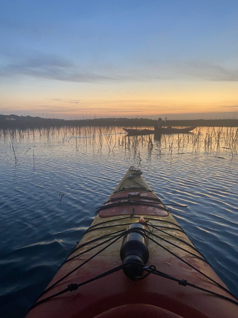 Tip of kayak on the water as the sun is almost set. Marsh grass in the background with another person on the kayak