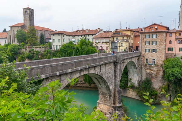 View from outside small towns in Italy showing a bridge over bright blue water with green vegetation and an old European looking village with terracotta rooftops across the bridge
