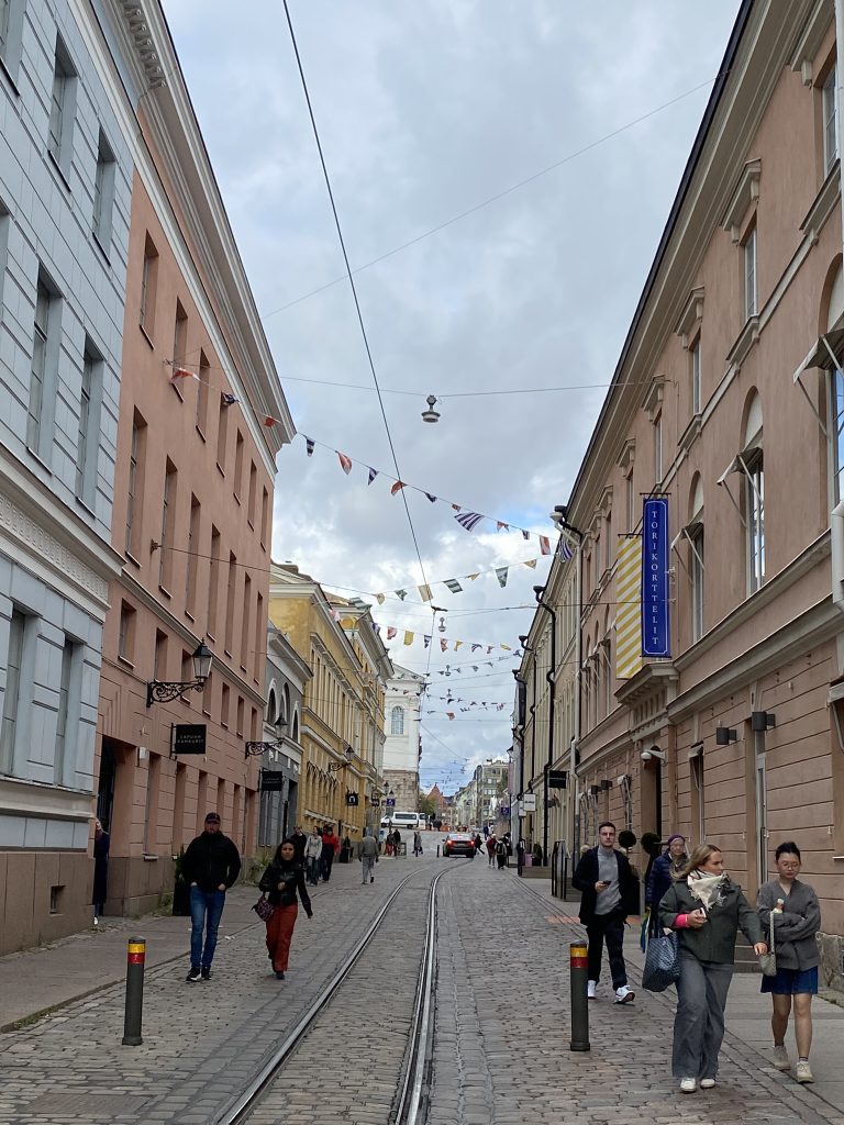 Main street in Helsinki with stores lining each side. The street has a streamer of flags ovehead as people walk down the street in romantic things to do in Helsinki