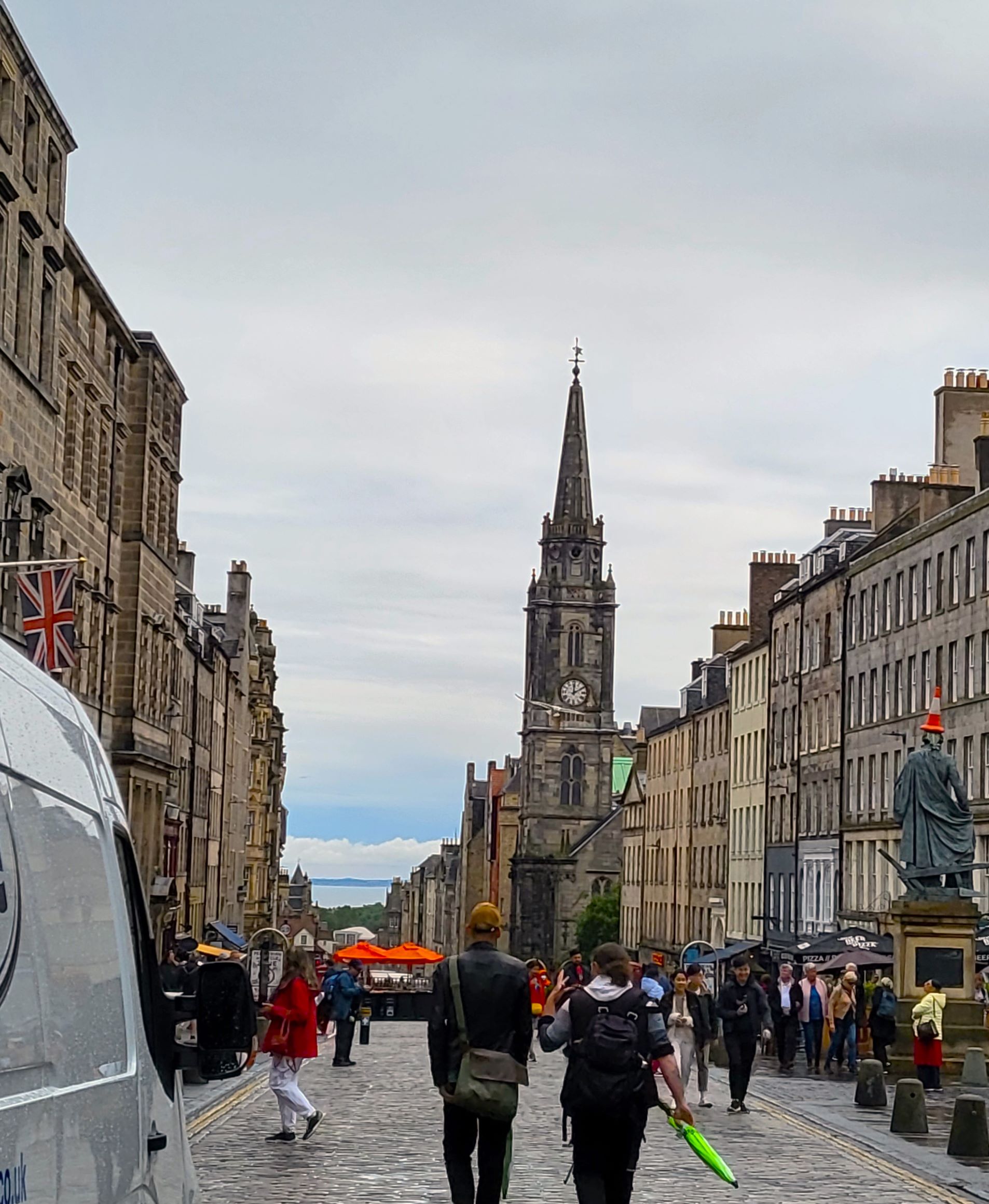 View down the street in Edinburgh on a dreary summer day. You can see the bell tower in the distance, shops on either side of the street as well as people walking down the center of the street in 21 cheap things to do in edinburgh