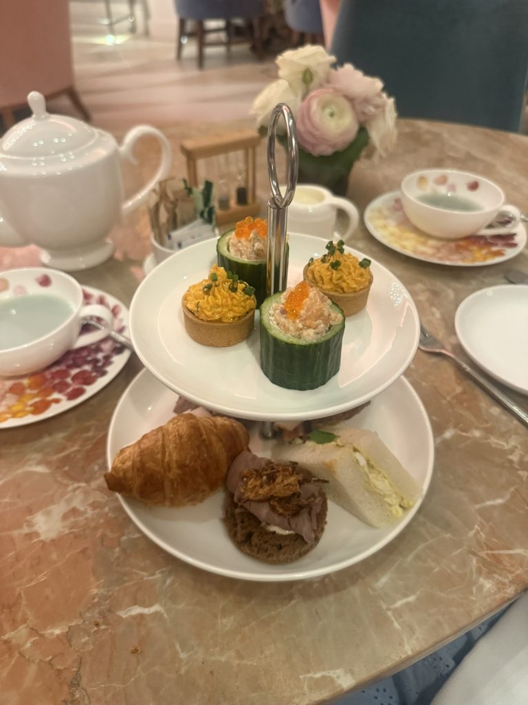 Two-tiered stand of savory afternoon tea bites, including cucumber cups topped with salmon roe, piped tartlets, a croissant, and tea sandwiches, served with a white teapot on a pink marble table at Camellias in Hotel Bennett