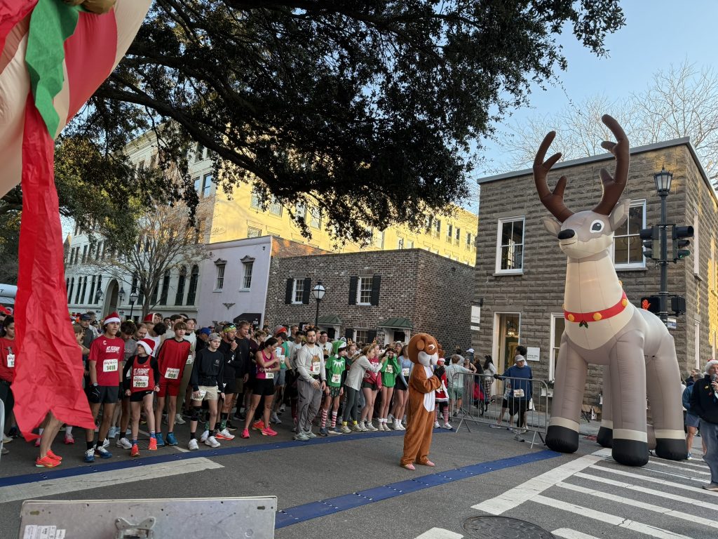 Runners in holiday costumes lined up at the Reindeer Run start line in downtown Charleston, with a giant inflatable reindeer and a costumed reindeer mascot in front of the crowd