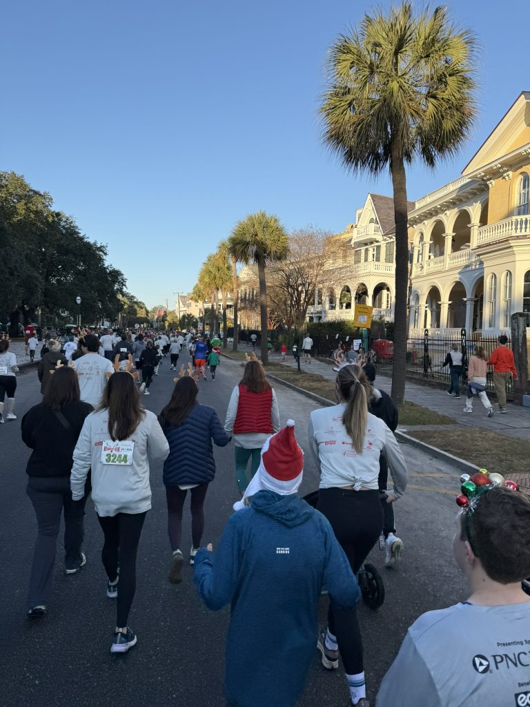 Runners and walkers in reindeer antlers and Santa hats during the Reindeer Run, passing palmetto trees and historic mansions along the Battery in downtown Charleston