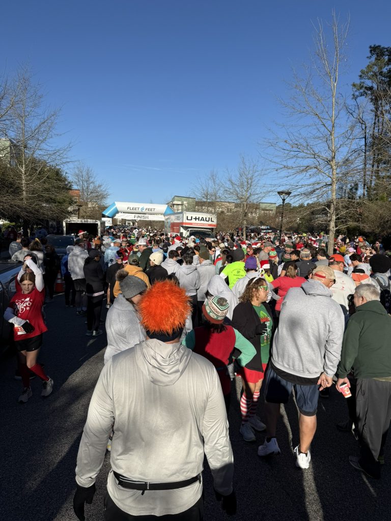 Crowd of runners in Santa hats, holiday sweaters and festive costumes gathered at the start of the Cocoa Cup 5K in Nexton, Summerville, with the start and finish arch in the background