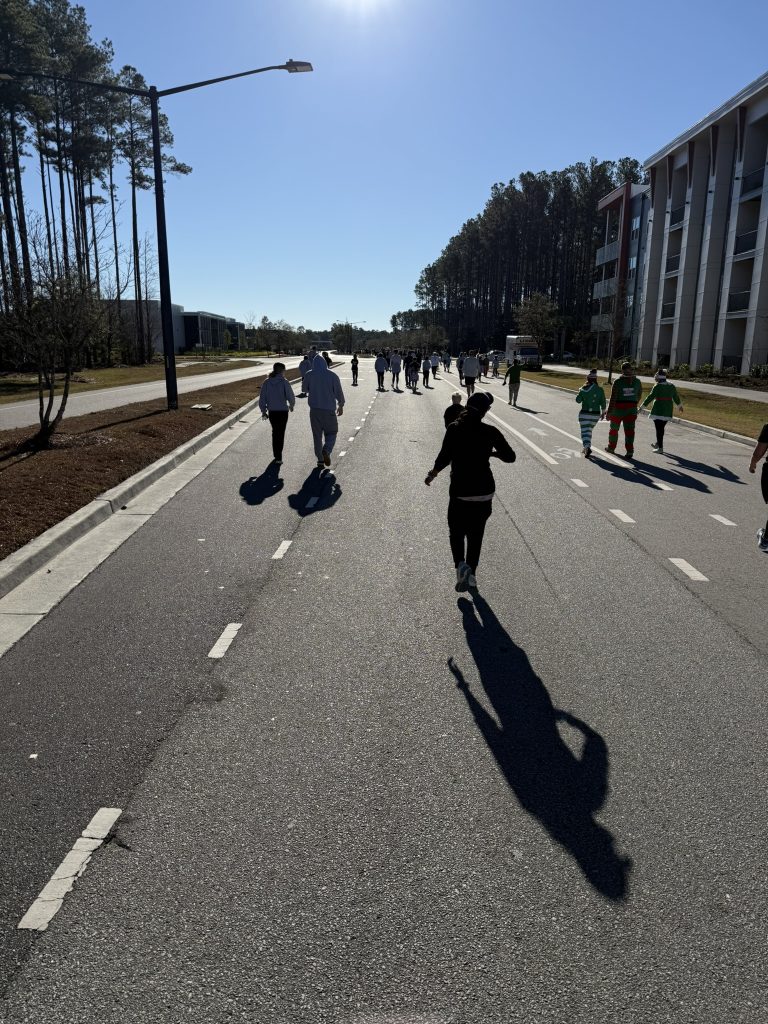 Runners, including a group dressed as elves, heading down a wide pine-lined road in Nexton on a sunny morning during the Cocoa Cup 5K in Summerville