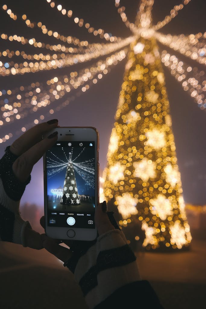 A person photographs a beautifully lit Christmas tree with a smartphone. Festive nighttime lights create a magical atmosphere with Christmas date ideas in Charleston, SC