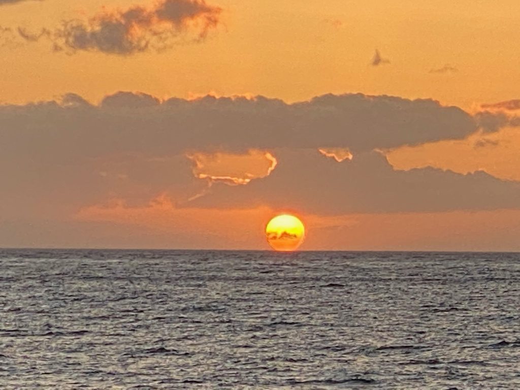 The sun just about to set beyond the ocean on a beautiful night with several clouds in the very colorful sky in maui vs the big island in Hawai'i