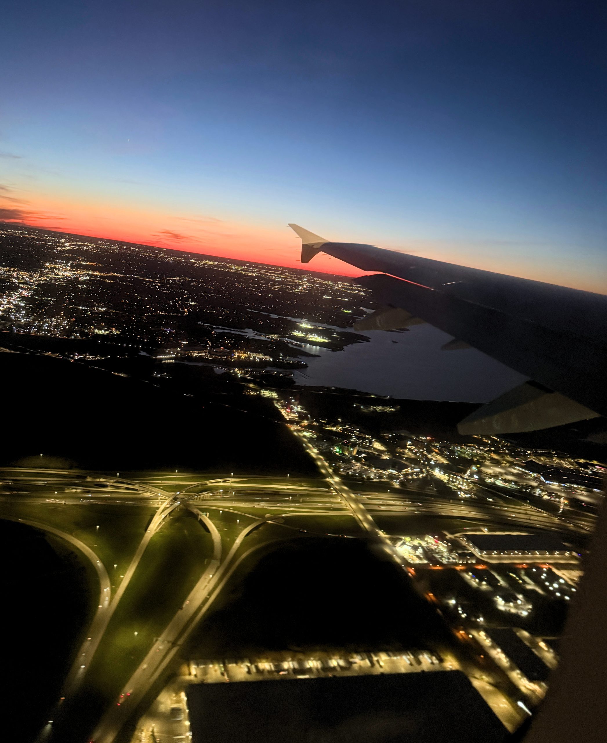 View out the window of an airplane. The sun is almost set on the horizon showing beautiful oranges and blues. You can see lights from a city below
