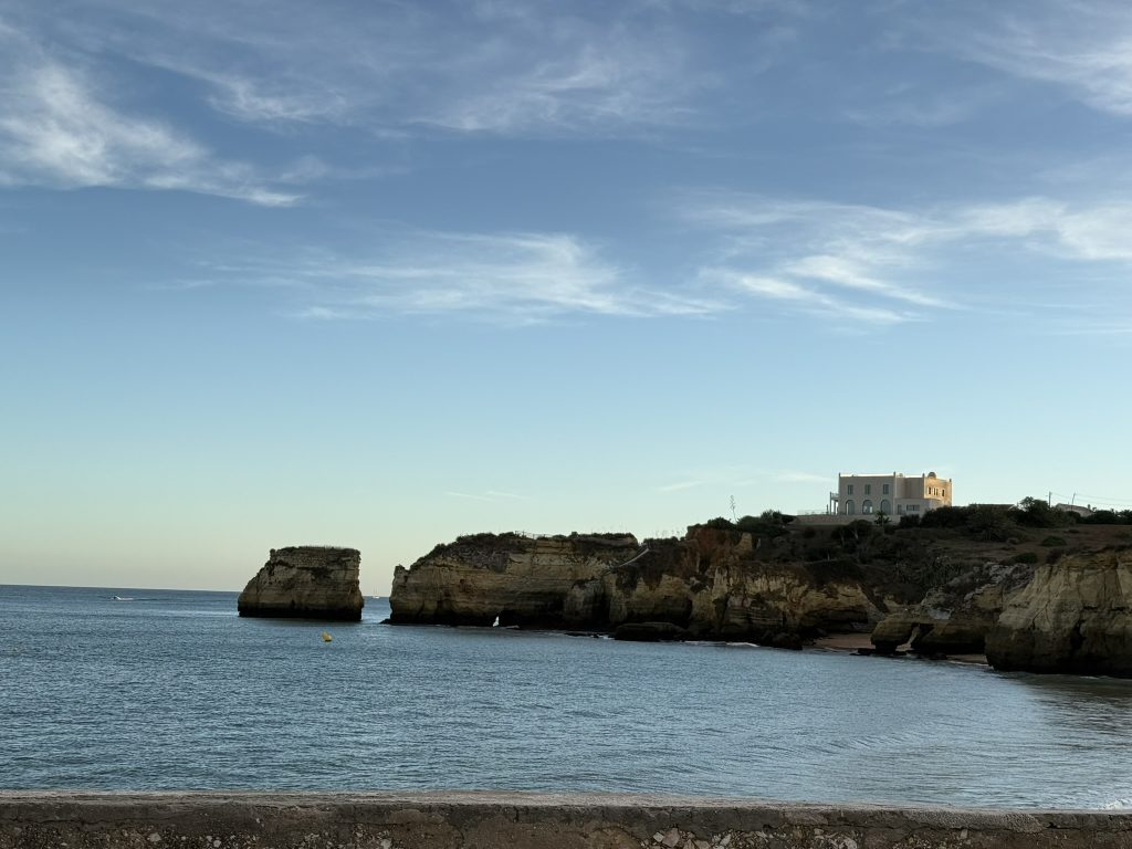 Shore line in Algarve, Portugal in the best Algarve resorts. A rocky cliff with a sole house on top is seen in the distance and the sandy beach is seen in the foreground