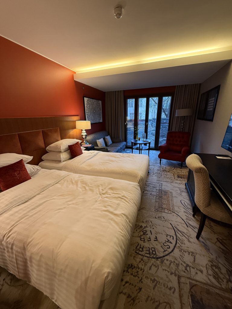 A room in the Telegraff Hotel in the Old Town part of Tallinn, Estonia. The room has a neat carpet with passport stamps, intimate lighting, 2 twin beds with nice bedding and fluffy pillows, a desk, tv, and sitting area with a couch and table, and a large opening window that looks out onto the medieval wall of the city.