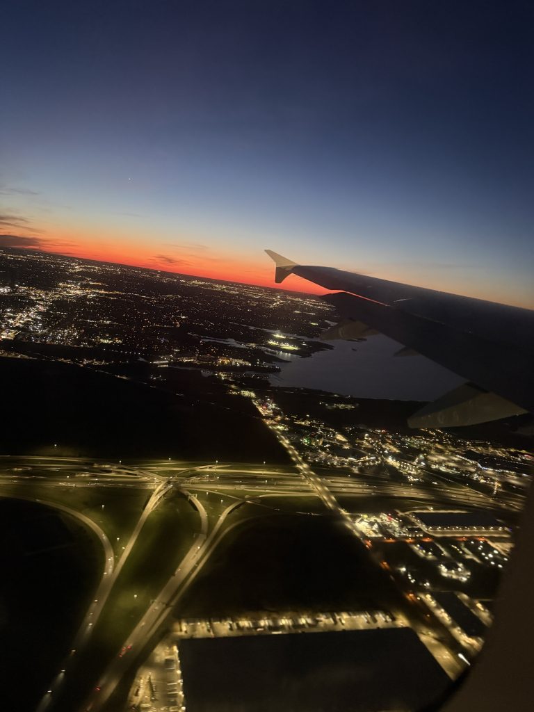 View out the window of an airplane. The sun is almost set on the horizon showing beautiful oranges and blues. You can see lights from a city below
