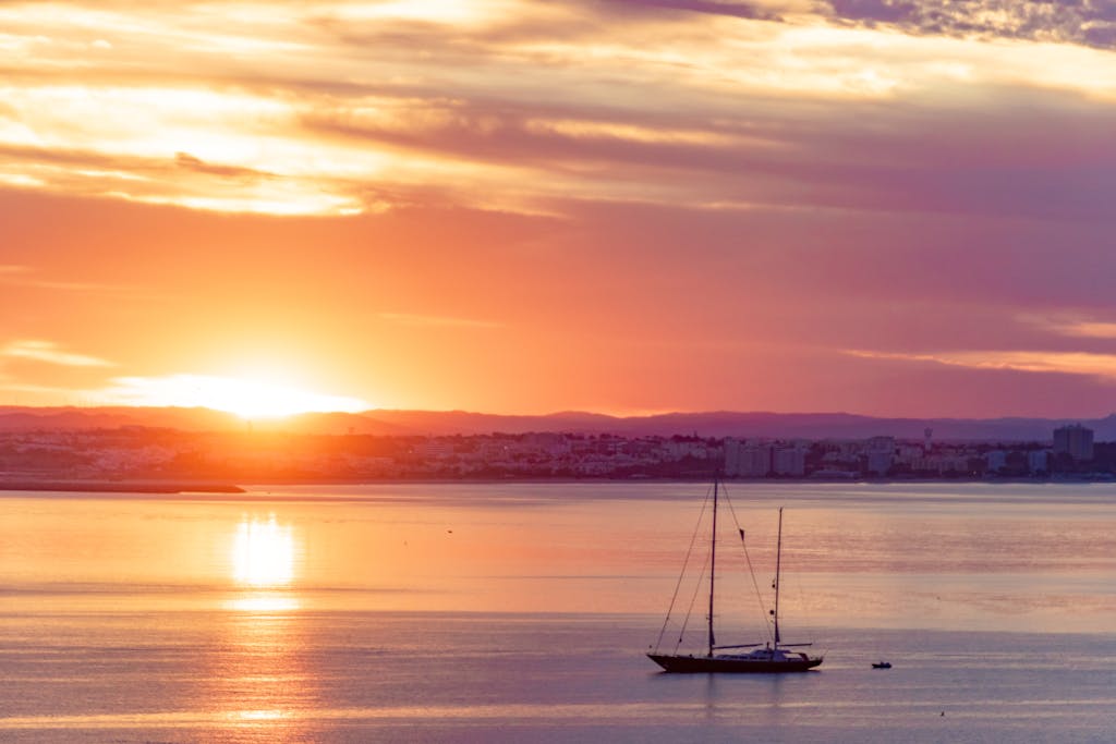 A serene sunset view over Lagos, Portugal in the best Algarve resorts,  with a silhouetted sailboat on the calm sea.