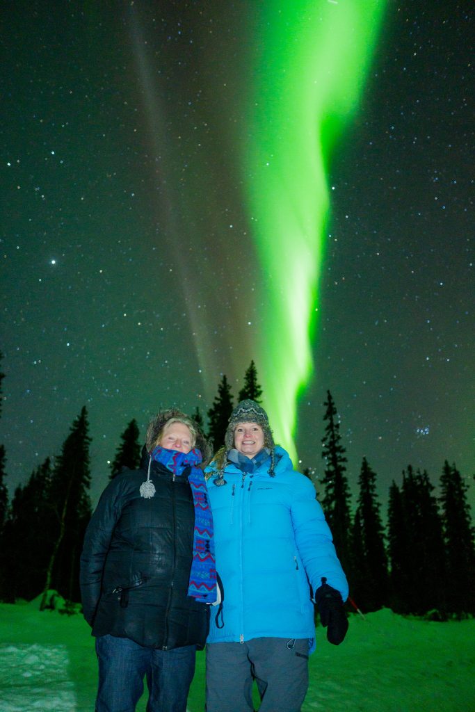 Casey Keller and her mother getting their picture taken with vibrant and electric green Northern Lights in the background in Swedish Lapland