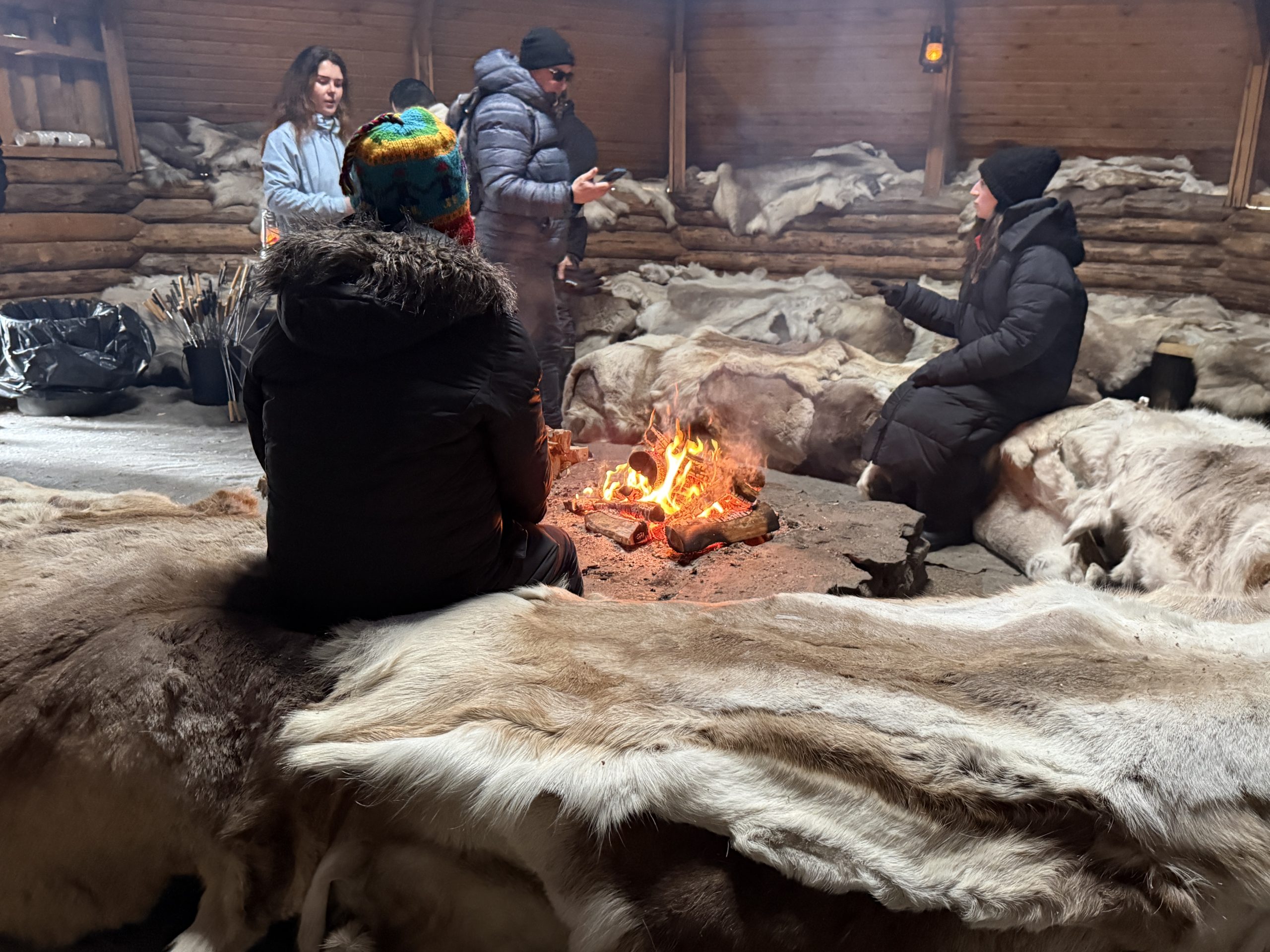 A traditional Sami hut with a fire in the center of the building warming the room. Guests are sitting on reindeer fur to stay warm.