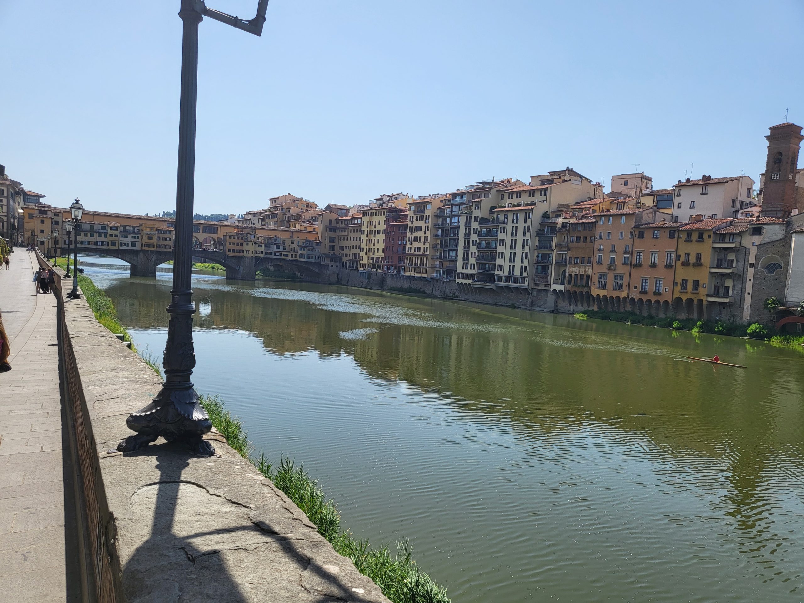 View on a sunny day down the river in hidden gems in Florence, Italy. Brightly colored houses and businesses can be seen across the river and an interesting looking bridge can be seen going over the bridge in the distance