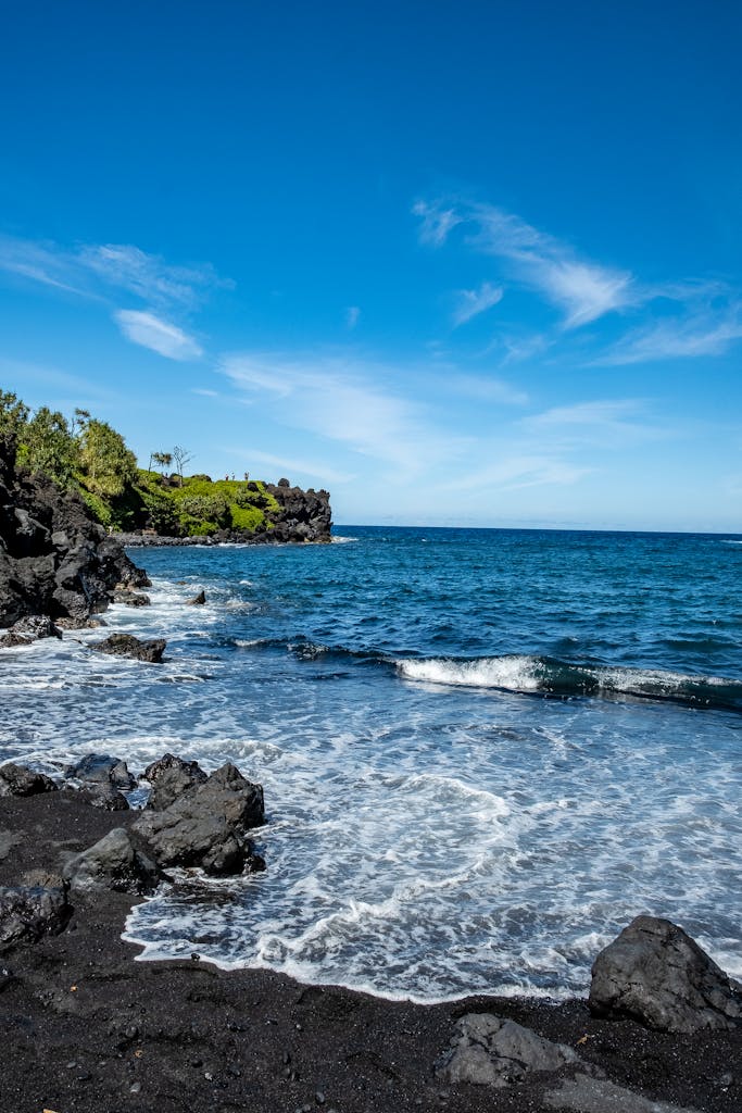 Stunning black sand beach with clear blue sky and ocean waves in Hana, Hawaii.