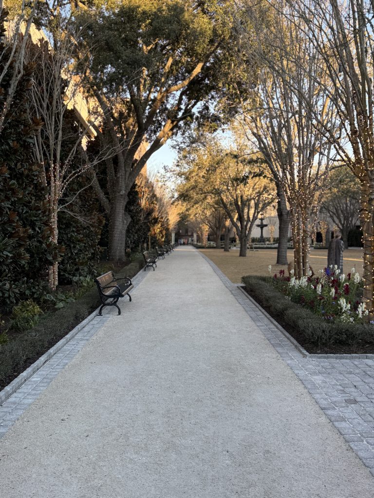 Walkway through American Gardens lined with trees and black benches. You can see flowers, green space and a fountain to the right, also within the park.