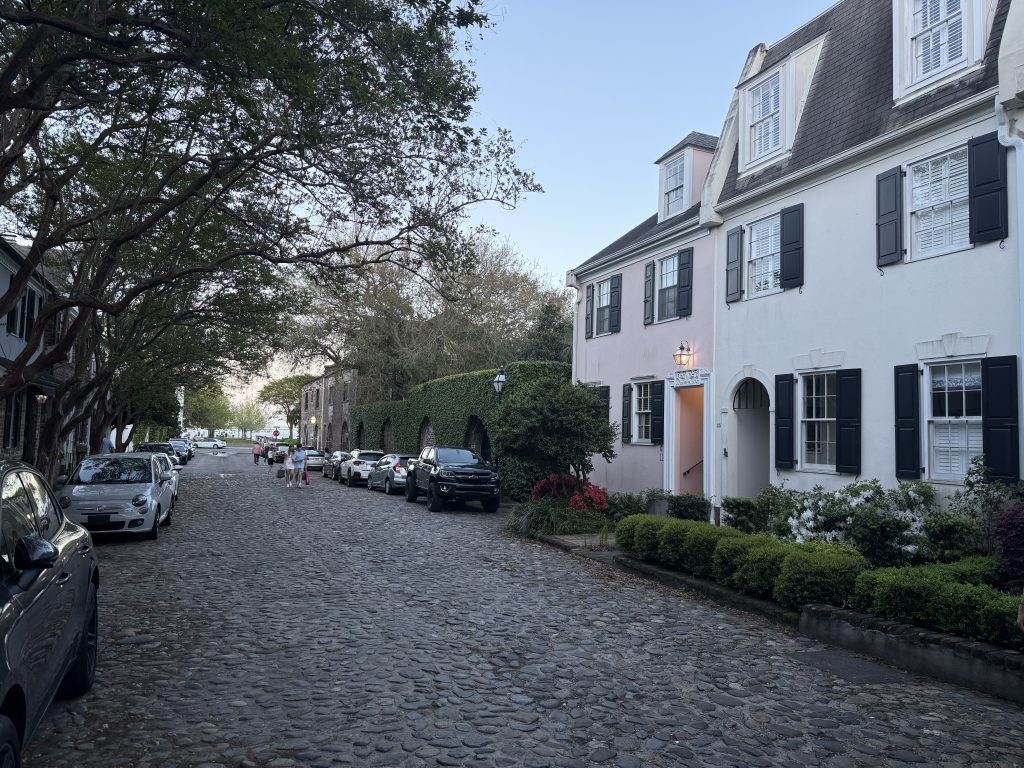 An old cobblestone street with colorful houses and cars lined up on the sides of the street in Charleston, South Carolina.