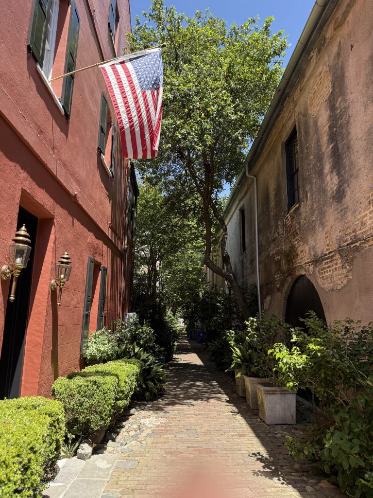 A view down Philadelphia Alley in Charleston, South Carolina. The alley has historic brick buildings on either side and is lined with green bushes. One building is adorned with an American flag.