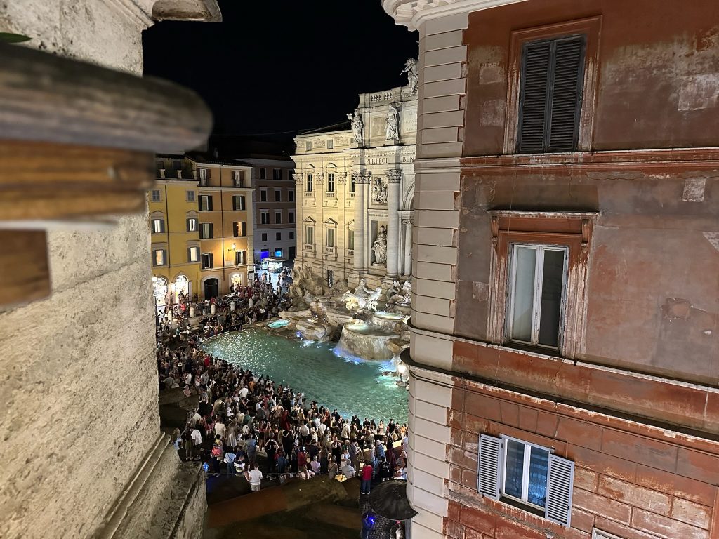 Tevi Fountain at night as seen from a cocktail bar across the plaza. Crowds of people are standing in front of the fountain