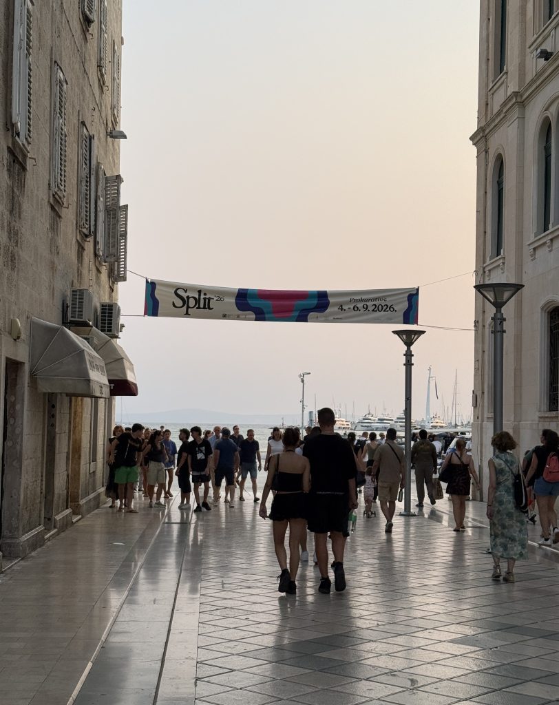 Crowds of people walk down a shiny paved street between pale stone buildings toward the waterfront in Split at dusk. A banner stretched across the street reads 'Split '26, Prokurative, 4.–6.9.2026' beside a bold pink, blue and purple wave design. Beyond the street, the sea, moored yachts and distant hills sit under a hazy peach sky, is Split worth visiting