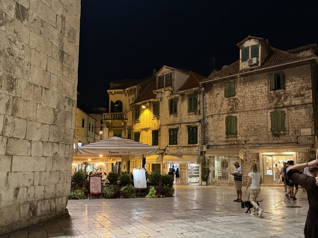 A stone-paved old-town square at night, lit by warm streetlights. A massive limestone wall fills the left side, and across the square stand weathered stone buildings with green shutters and terracotta roofs. A restaurant with a striped umbrella and potted plants has diners seated outside, and a few people stroll past shops, one walking a small black dog