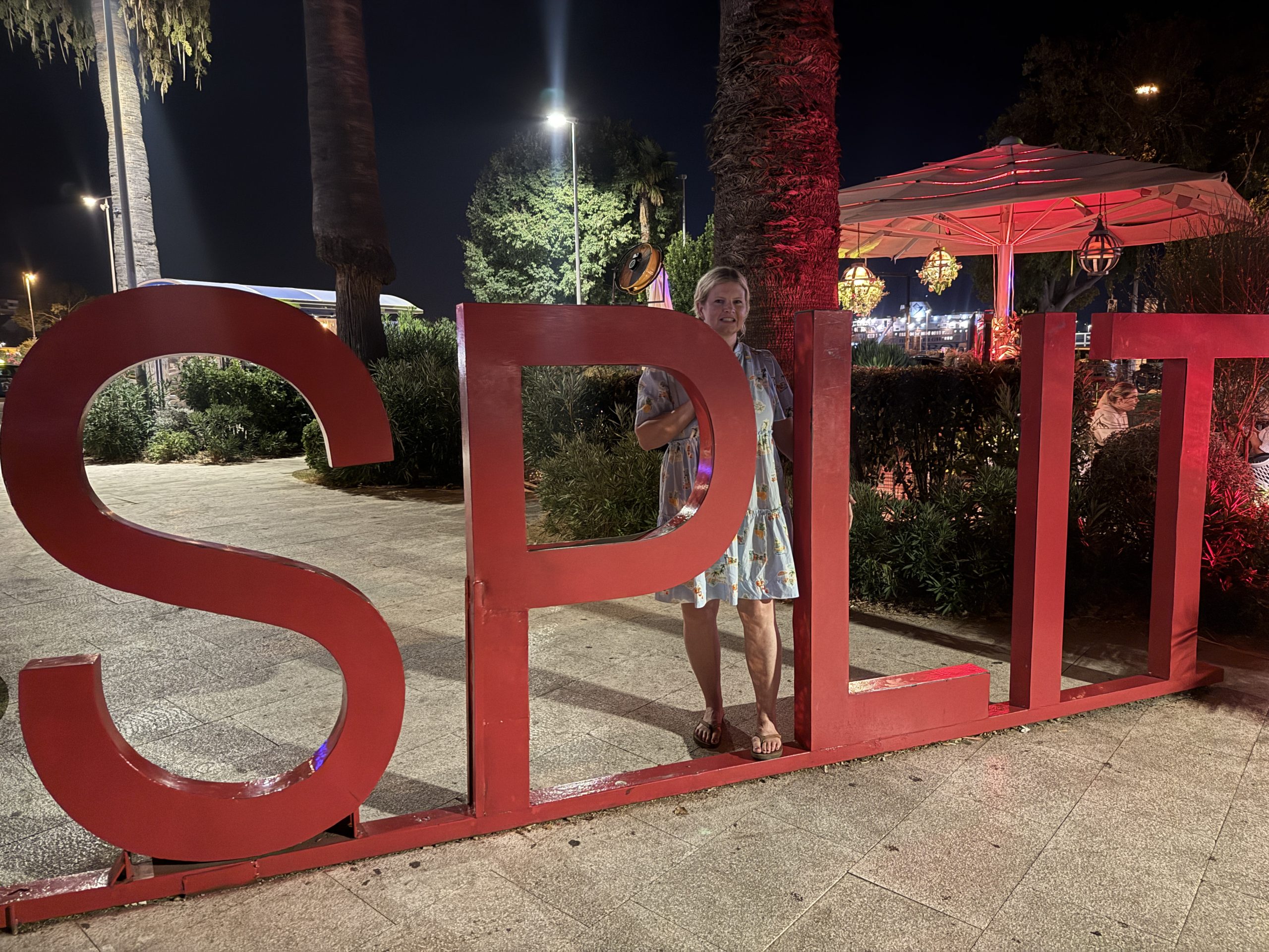 Casey of Wandering Everywhere smiling next to the large red "Split" letter sign at night in Split, Croatia, with palm trees and string lights in the background