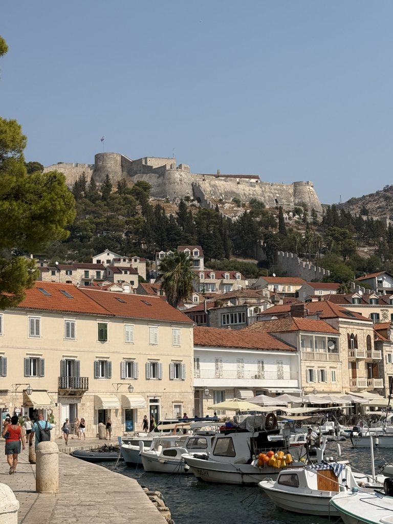 Hvar Town harbor with fishing boats, stone waterfront buildings, and the Fortica (Spanish Fortress) on the hill above