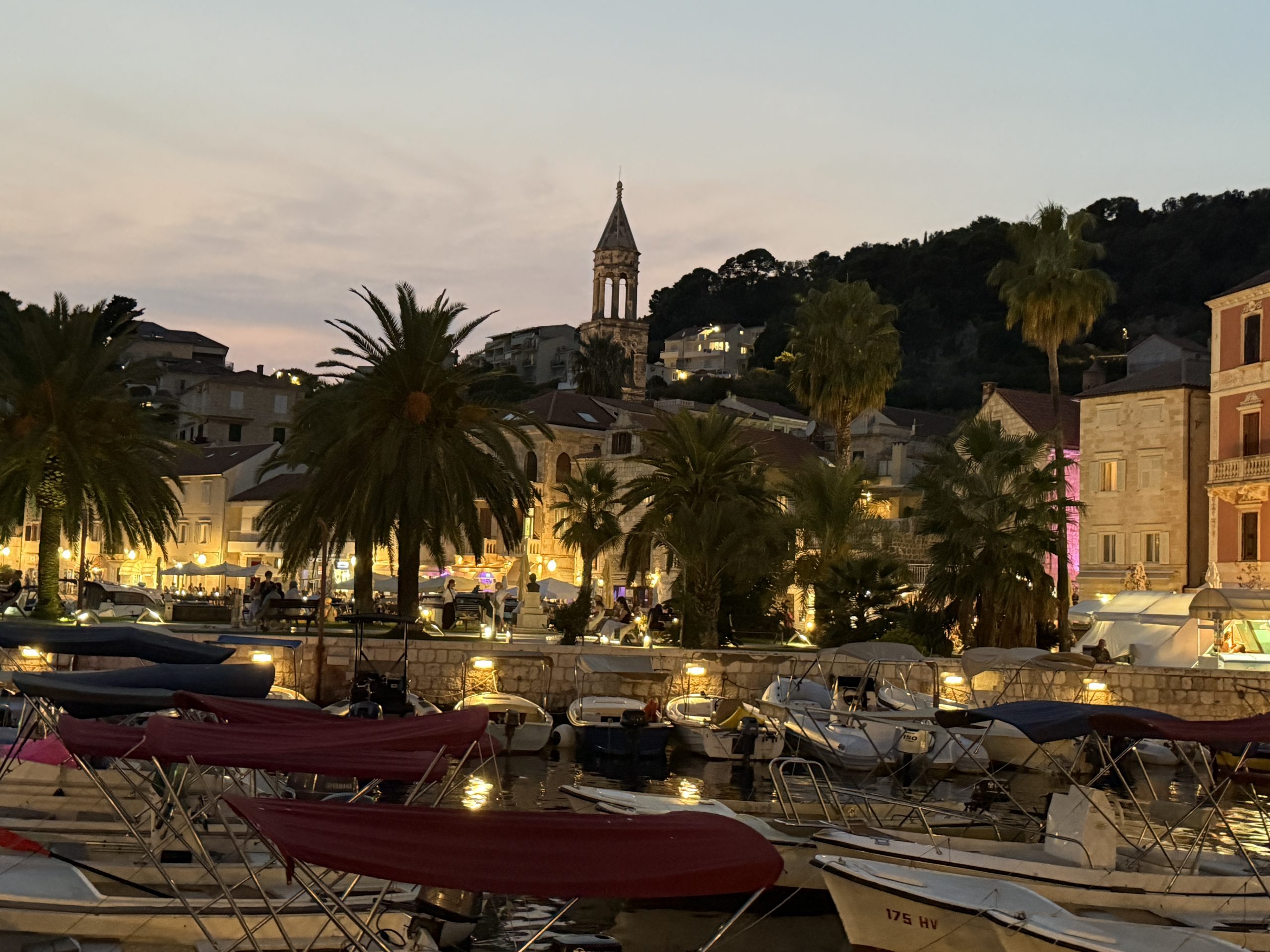 Hvar Town harbor at dusk, with small boats, glowing palm-lined waterfront cafés, and St. Stephen's Cathedral bell tower