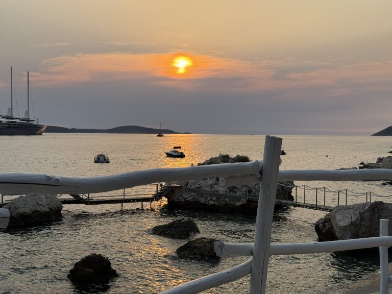 The sun setting through hazy orange clouds over the sea in Hvar Town, seen through a whitewashed rustic wooden railing. Below, rocks and a metal swimming platform sit at the water’s edge, small boats float in a path of golden light, and a three-masted ship is anchored by the Pakleni Islands on the horizon.