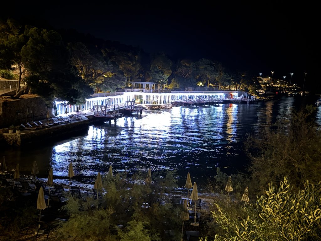 Waterfront beach club lit up white at night, with a long row of arched cabanas and canopied daybeds on wooden platforms over the sea. Their light reflects in rippling blue across a dark bay ringed by pine trees, with folded beach umbrellas on a pebble beach in the foreground.