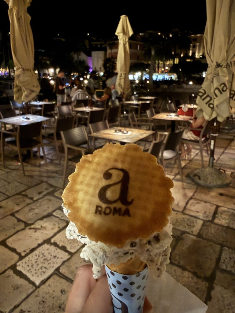 A hand holding a gelato cone with a speckled white scoop that looks like stracciatella, topped with a round wafer stamped with “a” and “ROMA.” Behind it is a café terrace on Hvar Town’s stone-paved waterfront at night, with tables, closed umbrellas and the glowing lights of the harbor.