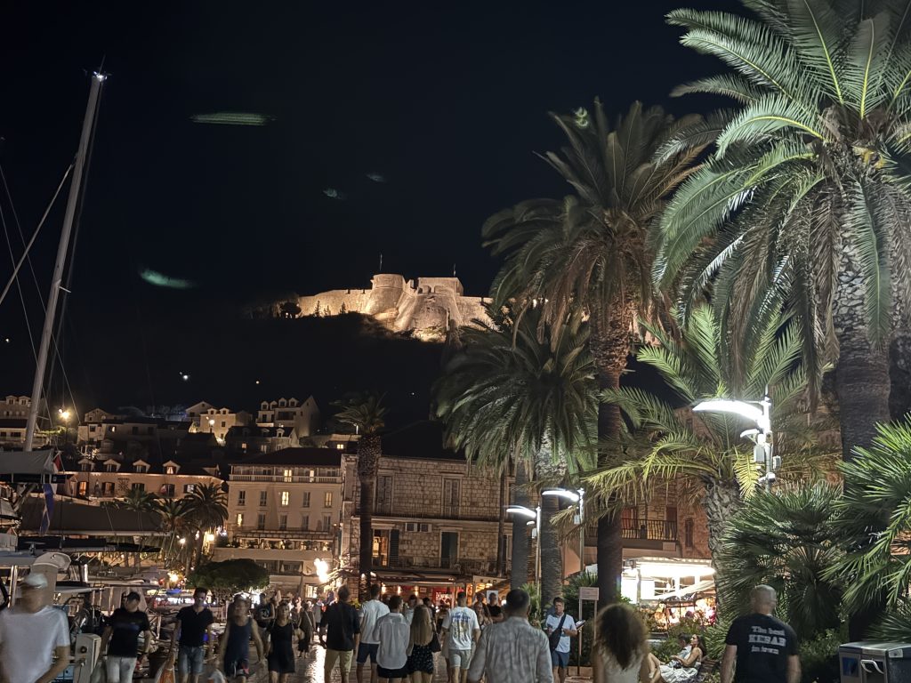 Hvar Town’s harbor promenade at night, crowded with people strolling under tall palm trees and streetlights past waterfront cafés and sailboats. The floodlit Spanish Fortress glows on the hill above the town.