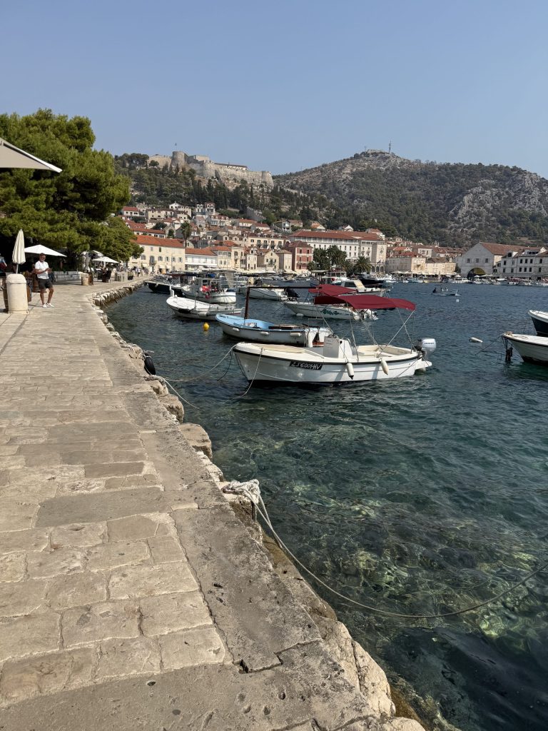 Limestone promenade along Hvar Town’s harbor, with small boats on clear turquoise water, terracotta-roofed Old Town buildings, and the hilltop Spanish Fortress