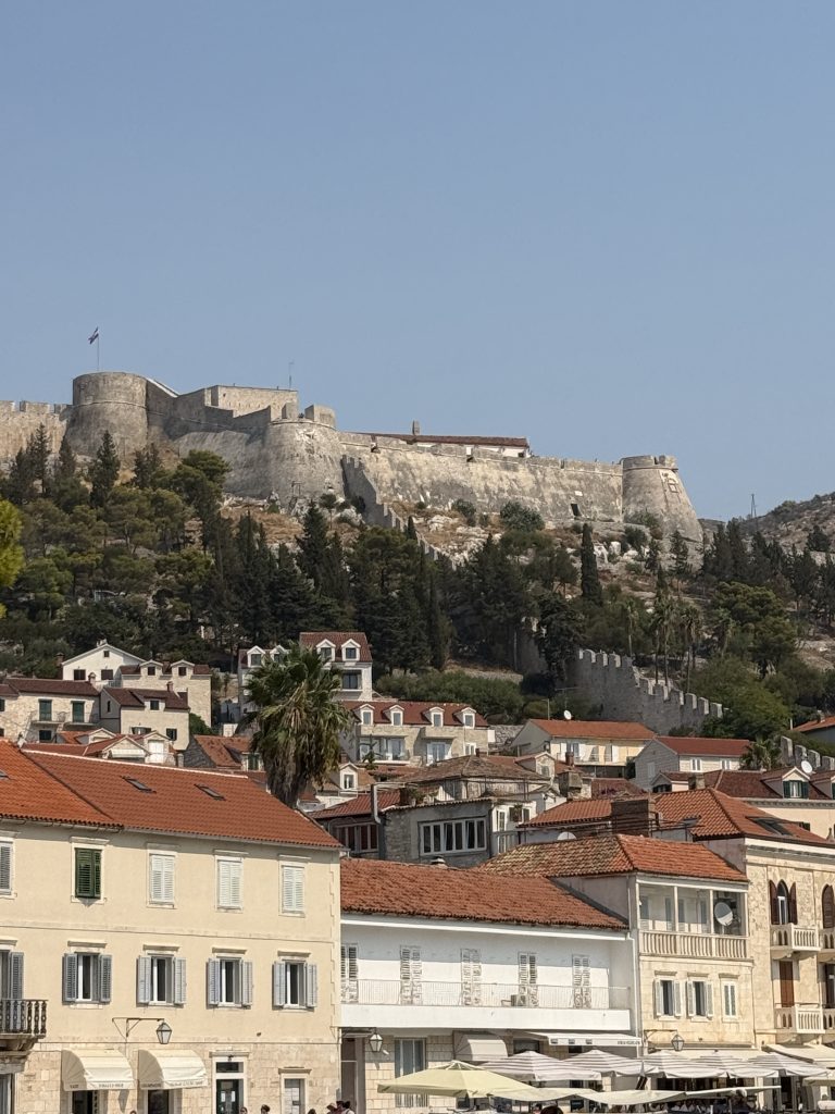 The Spanish Fortress in Hvar Town on a sunny day, its round stone bastions topped by a Croatian flag on a pine-covered hill. Crenellated medieval walls run down the slope to a cluster of red-tiled houses and cream-colored waterfront buildings with shuttered windows and café umbrellas.