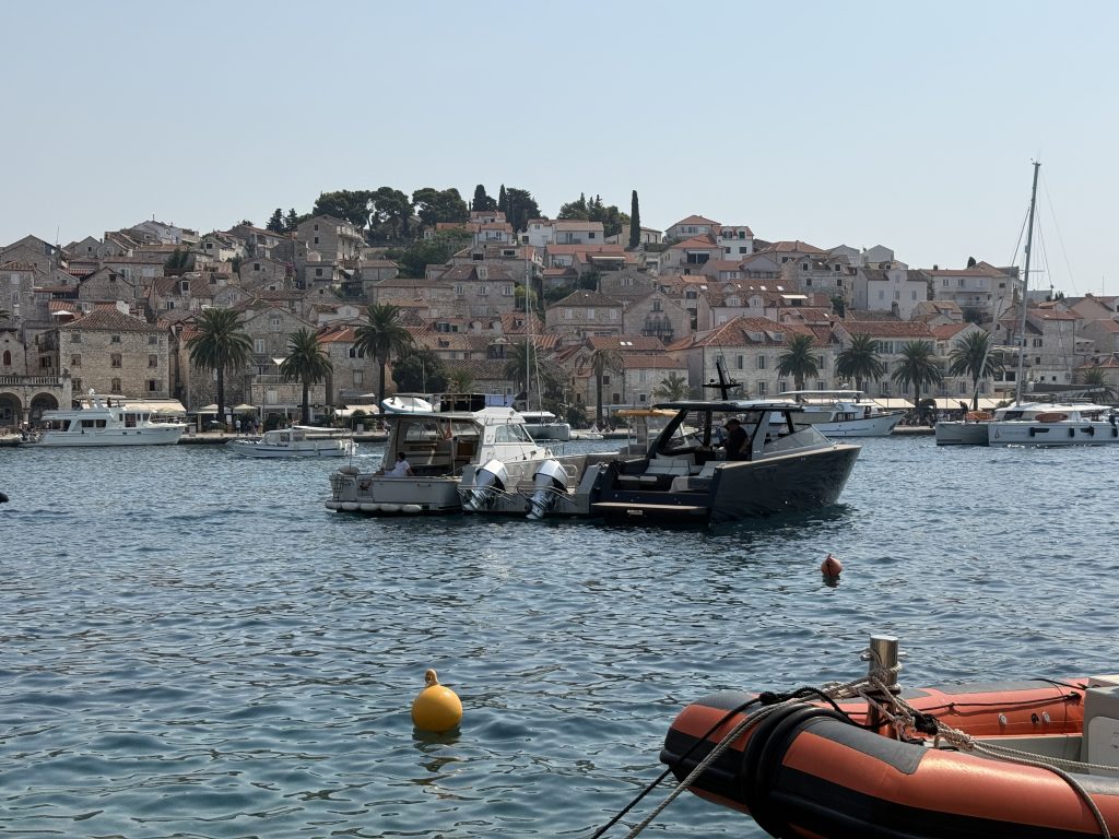View across Hvar Town’s harbor on a sunny day to a hillside of stone houses with red-tiled roofs above a palm-lined waterfront, with motorboats on the turquoise water and an orange inflatable boat moored in the foreground.