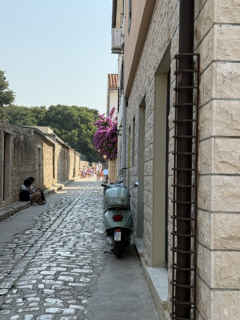 A narrow cobblestone lane in Hvar Town, just outside the pedestrian-only zone, with a mint-green Vespa parked against a stone building, purple bougainvillea spilling over a wall, and a group of children walking in the distance.