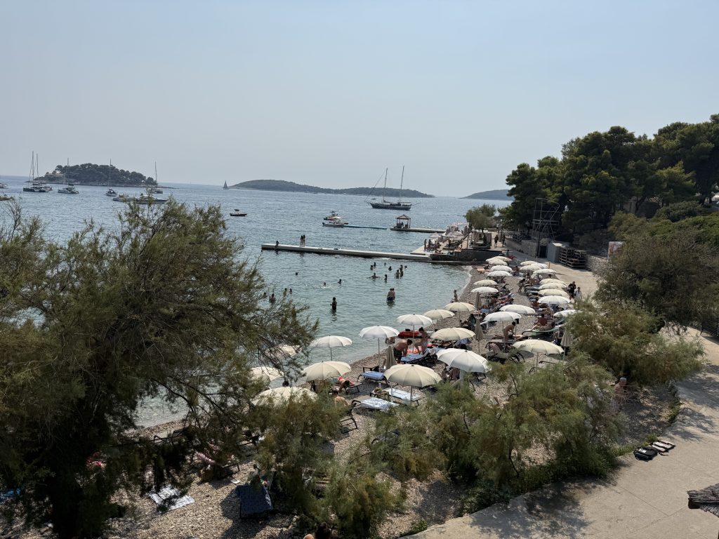 Pebble beach that was part of • Amfora Hvar Grand Beach Resort near Hvar Town lined with white umbrellas and sun loungers, with swimmers in clear water, a concrete swimming pier, and the Pakleni Islands offshore