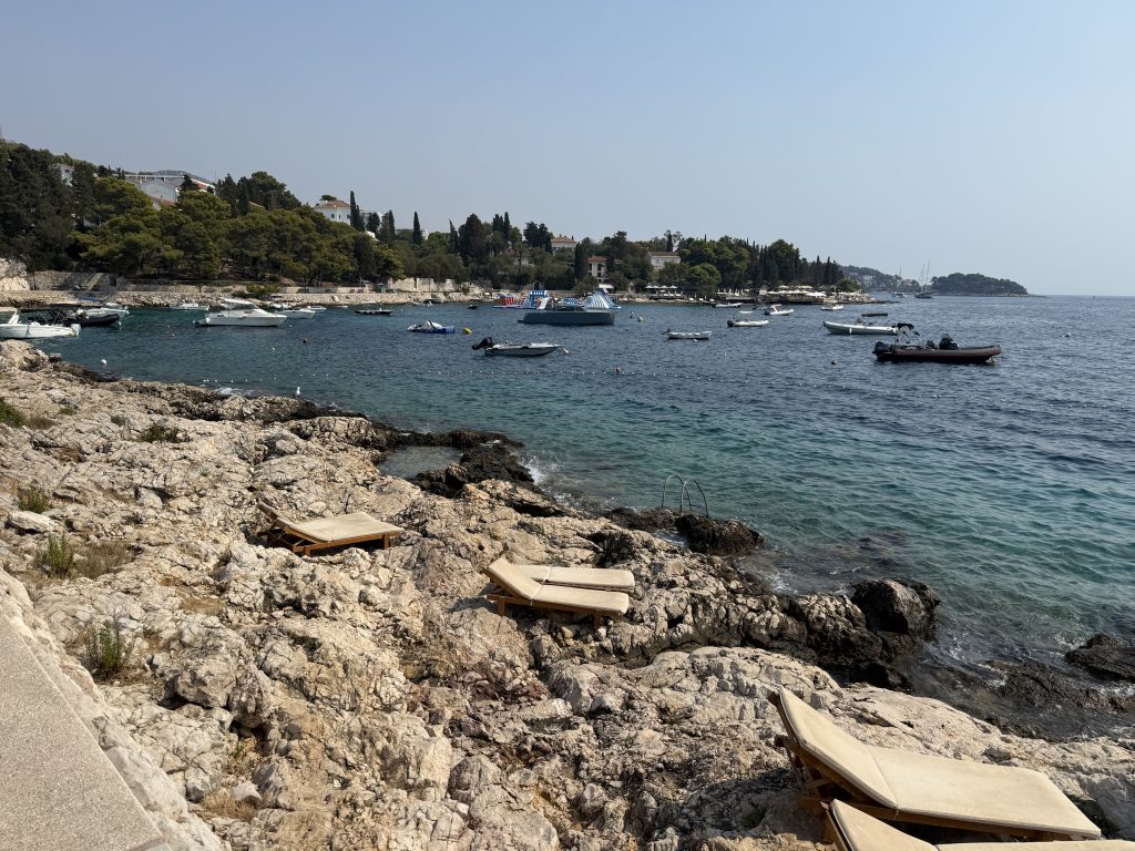 Rocky limestone shoreline along Hvar’s coastal path, with cushioned wooden sun loungers on the rocks, a metal swim ladder into clear turquoise water, and a cove of anchored boats, an inflatable water park, and a beach club beyond.