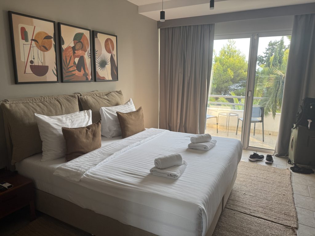 Guest room at Hotel Podstine in Hvar Town, with a king bed, folded towels, earth-toned wall art, and sliding doors to a private balcony shaded by pines and palms