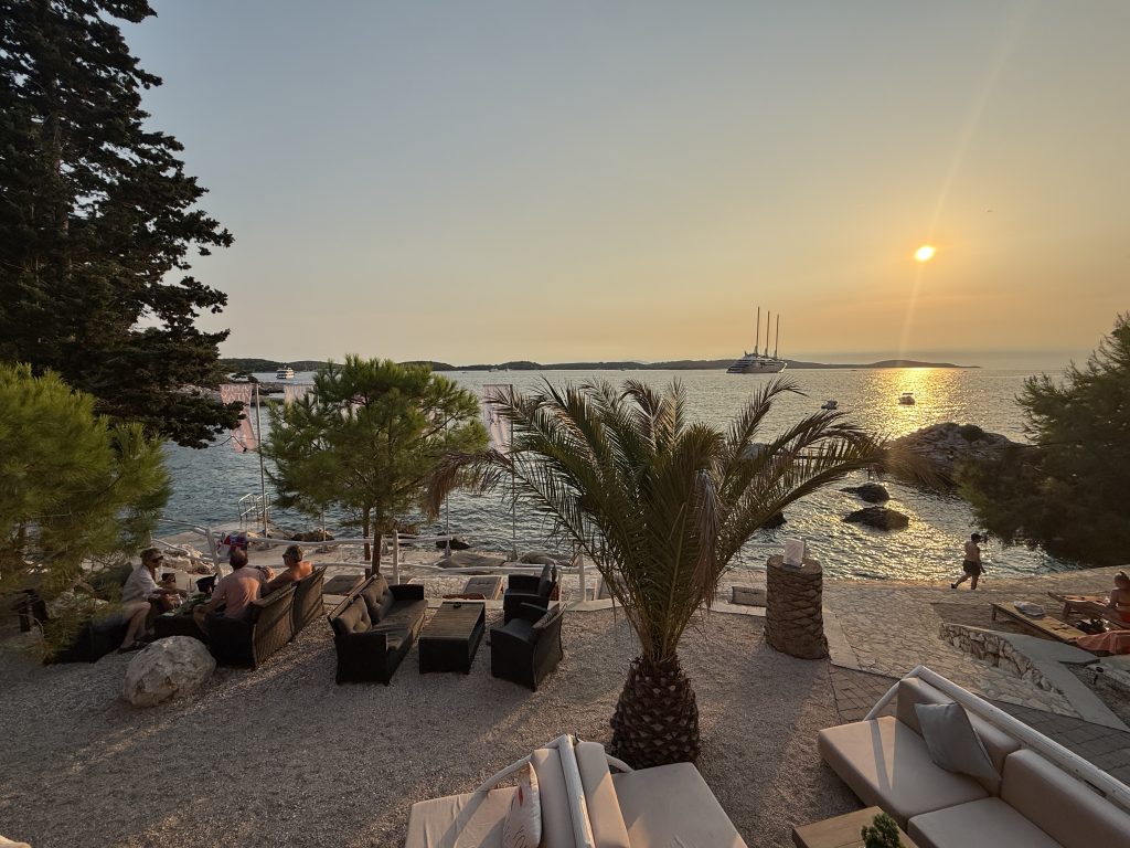 Sunset over the sea from a seaside lounge bar at Podsteine Hotel near Hvar Town, with people relaxing on outdoor sofas under palms and pines, and a three-masted ship anchored offshore
